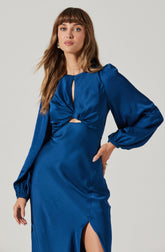A woman is wearing an elegant, royal blue satin dress with long, puffed sleeves that are gathered at the wrists. The top of the dress features a keyhole opening at the neckline, with fabric twisted into a knot just below the bust, creating a small cutout. The skirt has a thigh-high slit on the left side.
